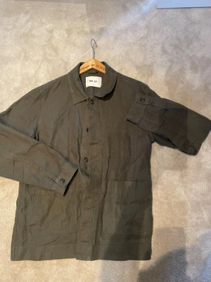 Olivgrön overshirt från NN07 - Snygg olivgrön overshirt från NN07 i storlek L. Tillverkad i ett lätt och luftigt material, troligen linne eller bomull. Klassisk krage, knäppning framtill och stora fickor. Perfekt lager-på-lager-plagg för en avslappnad stil.