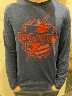 Blå långärmad topp Zadig & Voltaire - Snygg marinblå långärmad topp från Zadig & Voltaire med stort rött tryck framtill. T-shirten har rund halsringning och är tillverkad i mjukt bomullsmaterial. Perfekt för dig som gillar en cool och avslappnad stil. Väldigt sällsyn tröja, inte många har den. Skicket är mycket bra. Finns inget direkt tryck på storleken, men passar mig perfekt och jag är cirka 173cm lång. Pris är diskuterbart så var inte rädd att höra av dig!🙌 (Tröjan är äkta)