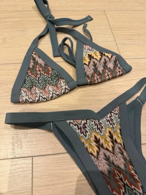 Mönstrad trekantsbikini med grå kant - Säljer en snygg trekantsbikini med grå kant och färgglatt mönster i grönt, rosa, gult och orange. Överdelen har knyt i nacken och ryggen, och underdelen har smala sidoband. Perfekt för strandhäng eller poolparty!