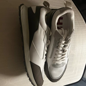 Vita sneakers från HUGO med bruna detaljer - Stilrena sneakers från HUGO i vitt med bruna och grå detaljer. Skorna har en chunky sula med HUGO-logga undertill, snörning och ovandel i en mix av läder och textil. Perfekta för en trendig och avslappnad look.