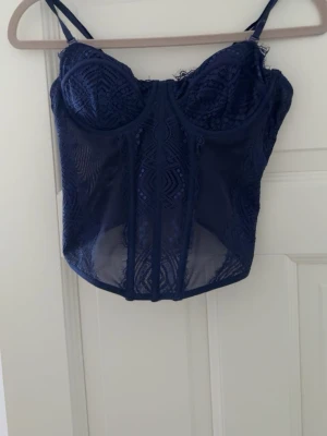 Mörkblå spetkorsett - Snygg mörkblå topp i spets med tunna axelband och vadderade kupor. Transparent mesh i midjan och dekorativa sömmar framtill ger en trendig look. Perfekt för dig som gillar detaljer och en feminin stil.
