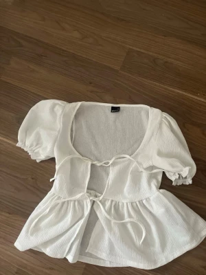 Vit blus med puffärm från Gina Tricot - Supersöt vit blus från Gina Tricot med puffärmar och två knyt framtill. Blusen har en luftig och lätt struktur, perfekt för varma dagar. Den har en feminin siluett med volang nedtill och djup urringning.