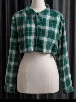 Grön rutig croppad skjorta - Grön och vit rutig croppad skjorta med klassisk krage och långa ärmar. Skjortan har knappar framtill och en loose passform som ger en avslappnad vibe. Perfekt för dig som vill ha en trendig och bekväm stil.