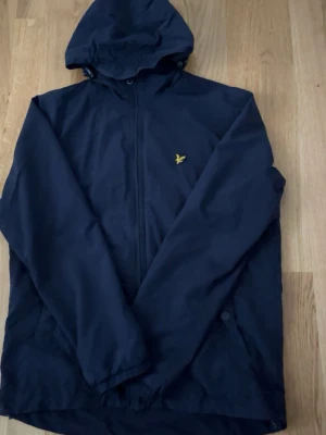 Svart vindjacka med huva Lyle & Scott - Snygg svart vindjacka från Lyle & Scott med gul logga på bröstet. Jackan har huva, dragkedja framtill och diskreta sidofickor. Perfekt för blåsiga dagar och enkel att matcha med olika outfits.