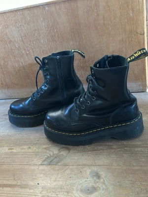 Svarta Dr. Martens Air Wair  - Säljer ett par klassiska svarta Dr. Martens kängor i skinn med grov sula och gul söm runt sulan. Kängorna har snörning, dragkedja på sidan och den ikoniska flärpen bak med logga. 