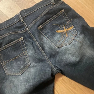 Mörkblå jeans från Cross Jeans - Säljer ett par mörkblå jeans från Cross Jeans. De har en cool broderad insekt på bakfickan och klassisk femficksdesign. Perfekta för vardagsbruk med en bekväm passform och snygg tvätt.