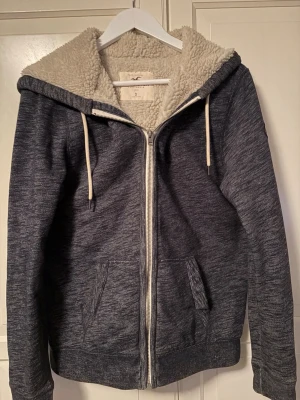 Mörkgrå hoodie från Hollister - Mysig mörkgrå hoodie från Hollister med dragkedja, två fickor fram och vit teddyfoder på insidan. Hoodien har huva med vita snören och är perfekt för kalla dagar. Materialet är mjukt och värmande, och designen är klassisk med en avslappnad vibe.