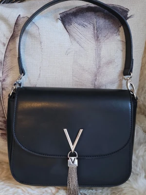 Valentino - Stilren svart handväska från Valentino i slätt läder med silverfärgad V-detalj och dekorativa kedjor framtill. Perfekt accessoar för dig som gillar klassisk men trendig design. Den är i fint skick 