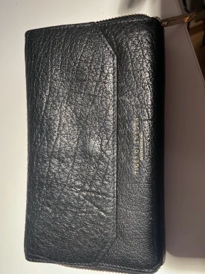Svart plånbok Tiger of Sweden läder - Snygg svart plånbok från Tiger of Sweden i äkta läder med struktur. Plånboken har guldfärgad dragkedja och präglad logga på framsidan. Klassisk rektangulär form och stilren design som känns både modern och tidlös.