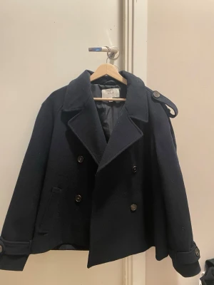 Mörkblå dubbelknäppt kappa VILA  - Säljer en mörkblå ullrock från vila med dubbelknäppning och stora slag. Rocken har axelklaffar, knappar vid ärmsluten och en klassisk, rak passform. Perfekt för dig som gillar stilrena och tidlösa plagg.