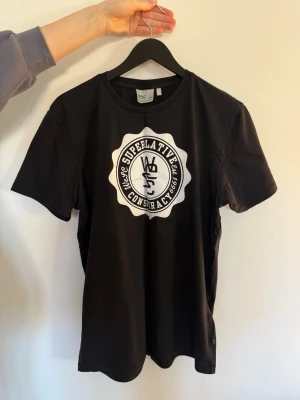 Svart Superlative Conspiracy t-shirt - Svart t-shirt från WeSC med stort vitt Superlative Conspiracy-tryck på bröstet. Klassisk rund halsringning och korta ärmar. Tillverkad i mjuk bomull för en skön känsla och enkel stil. Använd vid ett tillfälle.