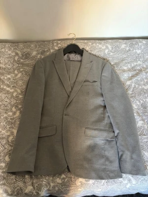 Ljusgrå kavaj med klassisk passform - Stilren ljusgrå kavaj med två framfickor med lock, bröstficka och smala slag. Kavajen har en enkelknäppning och är helfodrad. Perfekt för en dressad look och funkar till skjorta eller t-shirt.