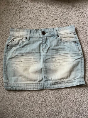 Ljusblå jeanskjol från Ellos - Lågmidjad ljusblå jeanskjol från Ellos med klassiska fickor fram och bak, snygga slitningar och detaljerad söm på bakfickorna. Kjolen har knapp och dragkedja framtill samt bälteshällor.