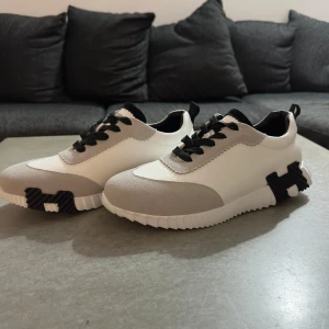 Vita och grå sneakers med svart sula - Säljer ett par fräscha sneakers i vitt och ljusgrått med svarta detaljer. Skorna har svart snörning och en unik sula med svart mönster på undersidan. Ovandelen är i syntet och mocka, perfekt för dig som gillar stilrena och moderna skor. Pris 999