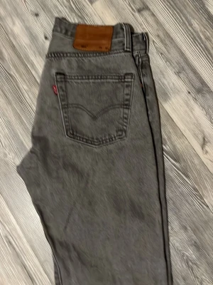 Grå Levi's raka jeans - Säljer ett par grå Levi's jeans med rak passform och klassisk femficksdesign. Jeansen har en brun läderpatch bak i midjan och den ikoniska röda Levi's-taggen på bakfickan. Tillverkade i slitstark denim, perfekta för en avslappnad och trendig look.