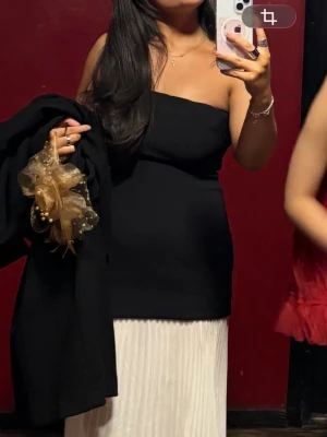 Svart och vit axelbandslös långklänning - Säljer en elegant axelbandslös långklänning med svart, figurnära överdel och vit, plisserad kjol. Klänningen har en stilren och modern look, perfekt för dig som vill sticka ut. Passformen är lite liten och kjolen faller snyggt längs benen.