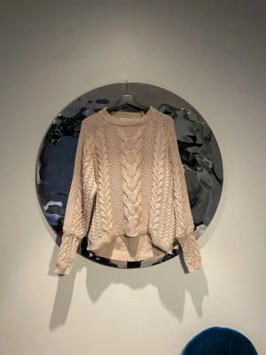 Beige kabelstickad tröja - Säljer en beige kabelstickad tröja med rund hals och ribbade muddar. Tröjan har ett snyggt flätmönster framtill och på ärmarna, samt en något längre bakdel för en avslappnad look. Perfekt för lager på lager under kyliga dagar.