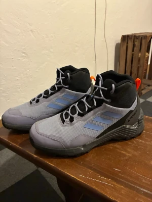 Grå Adidas Terrex kängor - Snygga grå kängor från Adidas Terrex med ljusblå detaljer och svarta partier. Skorna har en robust sula och mesh-material på ovansidan, samt orange dragflik bak. Perfekta för dig som gillar outdoor och sportig stil. Använda två gånger. Passade inte mig. Ursprungspris: 1399:-