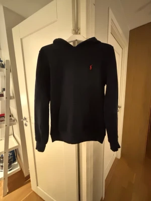 Mörkblå hoodie från Ralph Lauren - Snygg mörkblå hoodie från Ralph Lauren med klassisk huva och det ikoniska röda Polo-logotypen broderad på bröstet. Hoodien har en enkel och stilren design med ribbade muddar vid ärmar och nederkant. Perfekt för en avslappnad och trendig look.