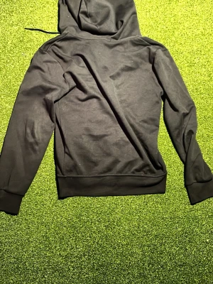 Svart hoodie från Polo Ralph Lauren - Svart hoodie från Polo Ralph Lauren med klassisk huva och ribbade muddar. Materialet är mjukt och bekvämt, perfekt för en avslappnad look. Diskret design utan tryck eller mönster, med dragsko i huvan för justering.