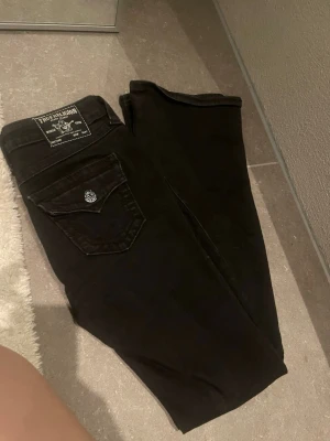 Svarta jeans från True Religion - Säljer ett par svarta jeans från True Religion med klassisk ficka och silverfärgad knappdetalj bak. Jeansen har botcut passform och är tillverkade i ett mjukt denimtyg som sitter snyggt på kroppen. Perfekta för dig som gillar stilrena och tidlösa jeans. Vet ej vilken modell dem är. storlek 27/32 