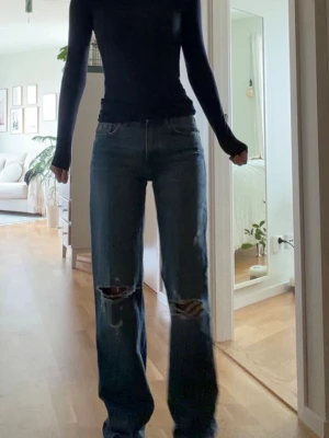Blå slitna raka jeans - Säljer ett par blå jeans med raka ben och stora slitningar över knäna. Jeansen är midrise och klassisk femficksmodell. Perfekta för en avslappnad och trendig look. Materialet är denim i bomull. 