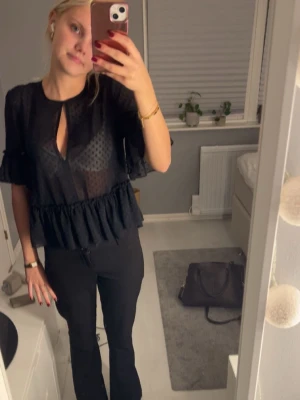 Svart volangblus med prickigt mönster - Trendig svart blus med volangdetaljer och diskret prickigt mönster. Blusen är lätt transparent med en snygg öppning vid halsen och har trekvartsärm. Perfekt för dig som gillar en chic och modern stil.