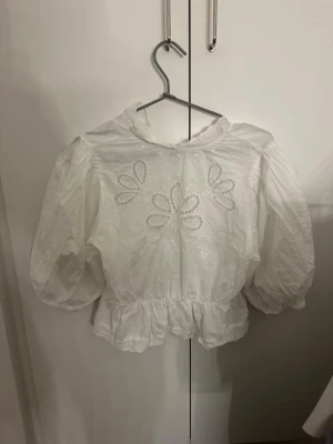Vit blus med puffärm och broderi - Söt vit blus med puffiga trekvartsärmar och broderade blommor framtill. Blusen har volangkant nedtill och spetsdetaljer vid hals och fåll. Tillverkad i tunn bomull, perfekt för en romantisk look. Från ginatricot i storlek Xs. Mycket nytt skick och andvänd endast en gång