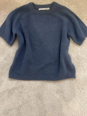 Blå stickad t-shirt från 157 - Mysig blå stickad t-shirt från 157 med korta ärmar och rund hals. Storlek 130 mem passar som 146/152 enligt mig, lager 157:s storlekar är aldrig helt korrekta😅 Tröjan har en enkel och stilren design med ribbade kanter vid hals, ärmslut och nederkant. Perfekt för dig som gillar en avslappnad men snygg look.