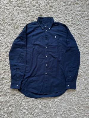 Mörkblå- Ralph Lauren - Klassisk mörkblå skjorta från Polo Ralph Lauren med vit broderad logga på bröstet. Skjortan har knappar hela vägen, button-down krage och långa ärmar. Tillverkad i mjuk bomull för en bekväm känsla. I princip helt oanvänd! 
