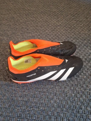 Adidas Predator fotbollsskor svart/orange - Säljer ett par Adidas Predator fotbollsskor i svart och orange med vita detaljer. Skorna har låg profil, strumpliknande ovandel och fasta dobbar för bra grepp på planen. Snygg design med klassiska tre ränder på sidan och Predator-tryck under sulan. Endast använd ett fåtal gånger. Strl UK 7.