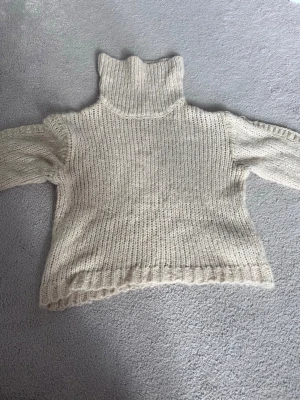 Beige stickad polotröja - Mysig beige stickad polotröja med bred ribbstickad krage och muddar. Tröjan har en avslappnad passform och är gjord i ett tjockare garn som ger en varm känsla. Perfekt för kalla dagar och enkel att styla till olika outfits.