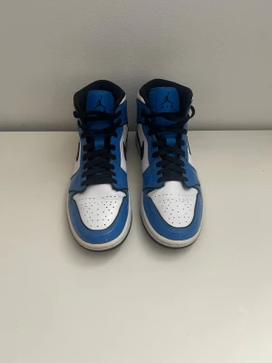 Nike Air Jordan 1 blå och vita sneakers - Säljer ett par Nike Air Jordan 1 sneakers i blått och vitt med svarta detaljer. Skorna har klassisk snörning, rund tå och är tillverkade i skinn med perforerad tåbox för extra ventilation. Perfekta för dig som vill ha en ikonisk och stilren sneaker.