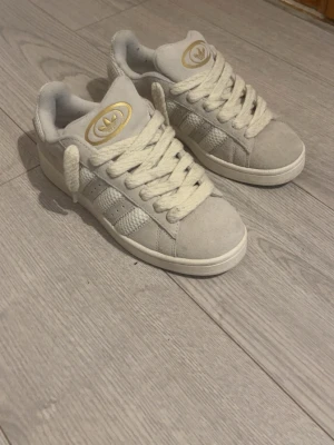 Adidas Campus  - Snygga vita Adidas sneakers med klassiska tre ränder på sidan och guldfärgad logga på plösen. Skorna har tjocka vita snören och ovandel i mocka och textil. Perfekta för dig som vill ha en clean och stilren look. Helt nya aldrig använda bara testat dom 1 gång 