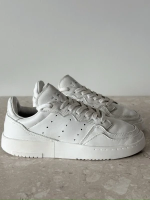 Vita Adidas sneakers i läder storlek 36  - Stilrena helvita sneakers från Adidas i mjukt läder med klassisk låg siluett. Skorna har perforerade detaljer på sidorna, diskret Adidas-logga på hälen och plös samt vit sula. Perfekta för en clean och modern look.