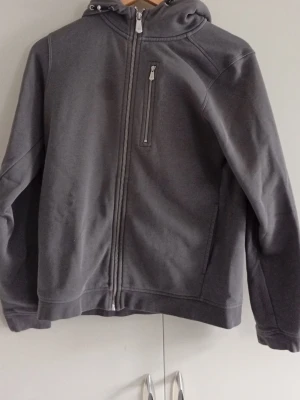 Sail Racing Kofta  - Snygg mörkgrå hoodie med hel dragkedja framtill och en bröstficka med dragkedja. Tröjan har huva med dragsko och långa ärmar. Perfekt för en avslappnad och stilren look. Materialet känns mjukt och bekvämt.