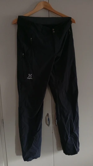 Svarta täckbyxor från Haglöfs - Säljer ett par svarta täckbyxor från Haglöfs med dragkedjor vid fickorna och resår vid bensluten. Byxorna har en enkel och stilren design, perfekta för kalla dagar utomhus. Materialet är slitstarkt och vattenavvisande.