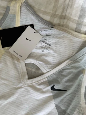 Nike Dri-FIT linne med grafiskt mönster - Sportigt linne från Nike med Dri-FIT-teknologi, perfekt för träning. Linnet har ett vitt basmaterial med ljusgrått och ljusgult grafiskt mönster, V-ringning och svart Nike-logga på bröstet. Tillverkat i ett lätt och ventilerande meshmaterial.