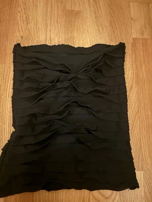 Svart bandeau tubtopp med rynk - Snygg svart bandeau tubtopp med rynkade detaljer och vågiga horisontella sömmar. Toppen är axelbandslös och har en tight passform som framhäver siluetten. Perfekt att styla med jeans eller kjol för en trendig look.