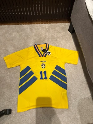 Sverige hemmaställ vm 94 - Brolin #11  - Klassisk gul Sverige fotbollströja från Adidas med blå detaljer, nummer 11 och Brolin på ryggen. Tröjan har V-ringad krage med blå och gula ränder samt det svenska landslagets emblem på bröstet. Perfekt för dig som älskar retro och landslagskänsla.