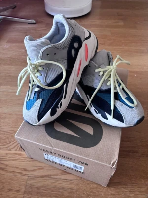 Yeezy Boost 700 Wave Runner sneakers - Säljer ett par Yeezy Boost 700 Wave Runner sneakers med grå, blå och svarta paneler, gula skosnören och vita samt orange detaljer på sulan. Skorna har en chunky siluett och är tillverkade i mesh, mocka och läder för en riktigt modern vibe.