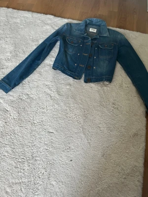 Klassisk blå jeansjacka - Snygg blå jeansjacka med kortare passform och klassiska bruna knappar. Jackan har två bröstfickor med lock och knapp samt långa ärmar. Perfekt att slänga över en hoodie eller t-shirt för en cool look.