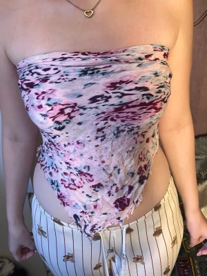 Blommig bandeau tubtopp i rosa - Trendig bandeau tubtopp i ljusrosa med blommigt mönster i vinrött och mörkblått. Toppen är axelbandslös och knyts i ryggen för en snygg och avslappnad look. Perfekt till sommaren och passar till både jeans och kjol.