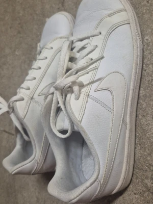 Vita låga sneakers från Nike - Säljer ett par klassiska vita sneakers med lågt skaft från Nike. Skorna har snörning och är tillverkade i slätt läderliknande material med vit sula. Perfekta för en clean och stilren look. Skorna är använda en gång i någon timma cirka bara då dom var för stora för mig, har legat i källaren sedan dess.