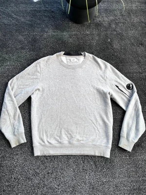 C.P. Company sweatshirt  - Snygg grå sweatshirt från C.P. Company med rund halsringning och ribbade muddar. Tröjan har en ikonisk linsdetalj på ena ärmen och är tillverkad i mjuk bomull. Perfekt för en clean och avslappnad stil. Skick 9/10, storlek L. Pris går att diskutera✅
