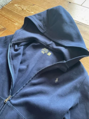 Mörkblå hoodie från Polo Ralph Lauren - Snygg mörkblå hoodie med dragkedja från Polo Ralph Lauren. Klassisk design med huva, fickor framtill och den ikoniska broderade loggan på bröstet. Tillverkad i mjukt bomullsmaterial som är skönt att ha på sig. Perfekt för en chill och avslappnad stil.