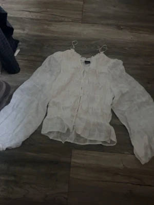 Vit blus med puffärm från Gina Tricot - Superfin vit blus från Gina Tricot med puffiga långärmar och smockdetaljer. Blusen har knappar framtill och tunna axelband för en trendig look. Tillverkad i ett lätt och luftigt tyg som ger en romantisk känsla.
