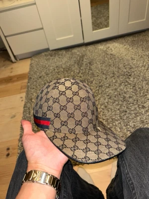 Beige keps - Säljer en beige keps med klassiskt GG-mönster över hela kepsen och en snygg röd-blå rand på sidan. Justerbar rem bak för perfekt passform. Perfekt accessoar för dig som vill sticka ut med en ikonisk look. Den har slitage märken som t:ex en repa vid toppen av skärmen på vänstersidan av kepsen. den är absolut i acceptabelt skick men har märken användning men det får man såklart för dehär priset