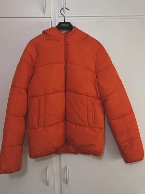 Orange pufferjacka med huva - Snygg orange pufferjacka med huva och dragkedja framtill. Jackan har två stora fickor på sidorna och quiltad design som ger en cool look. Perfekt för kalla dagar när du vill hålla värmen och ändå se trendig ut.