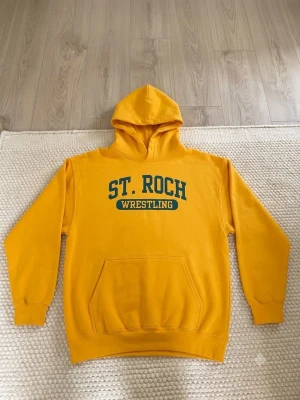 Gul hoodie ST. ROCH Wrestling - Gul hoodie med huva och magficka, tryck med texten 'ST. ROCH WRESTLING' i grönt och gult på bröstet. Tillverkad i 100% bomull, perfekt för dig som gillar sportig och avslappnad stil. Klassisk passform och ribbade muddar vid ärmar och nederkant.